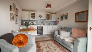 A modern living room with a grey sofa, a dining table with chairs, and a kitchen area with white cabinets and a clock. - The Linhay (Barnstaple)