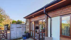 A wooden cabin with a stone wall, a sliding glass door, and a patio area with a chair and a blue trash bin. - The Linhay (Barnstaple)