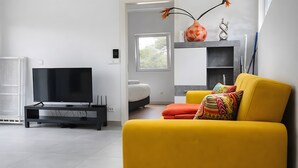 En moderne stue med et fladskærms-tv, en gul sofa, en hvid seng og et vindue med naturligt lys.