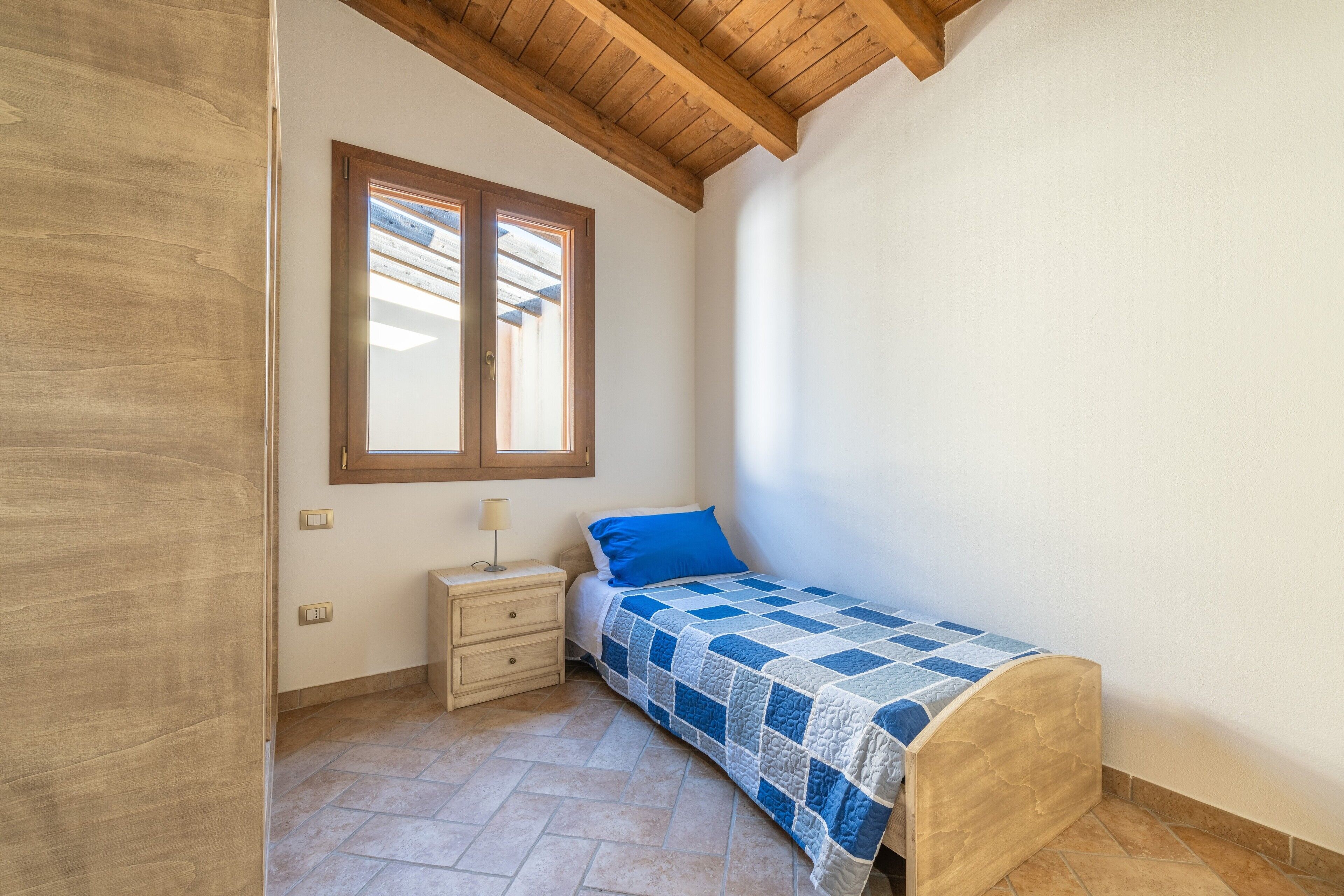 Ein Schlafzimmer mit einem Bett, einem Nachttisch, einem Fenster und einer Holzbalkendecke.