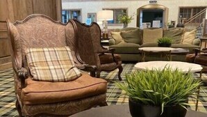 Un vestíbulo con piso a cuadros, una mesa redonda con un periódico y un sillón marrón con cojín de cuadros.