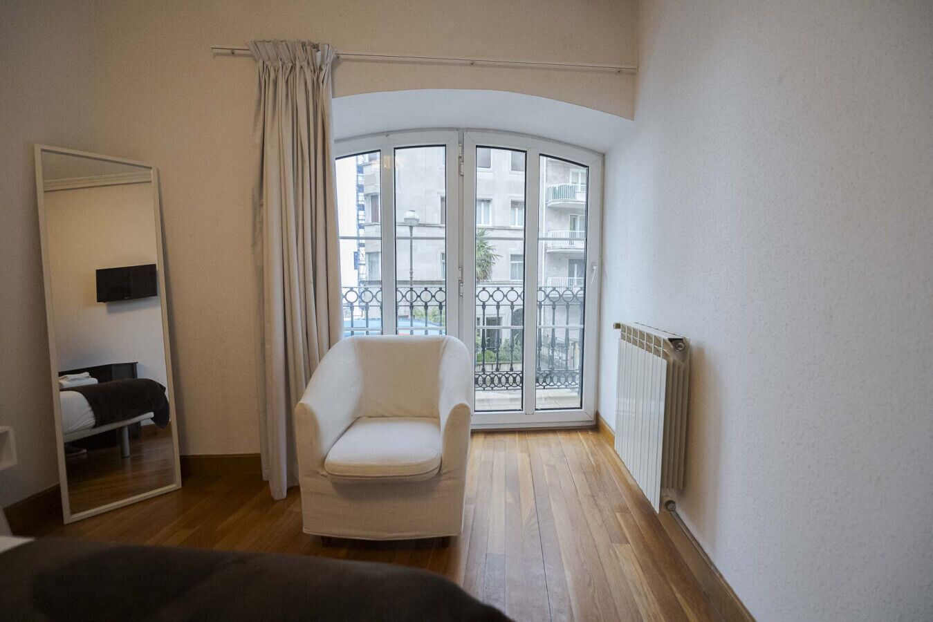 Ein Zimmer mit weißem Sessel, einem großen Spiegel, einer Heizung und Blick auf einen Balkon mit Geländer.