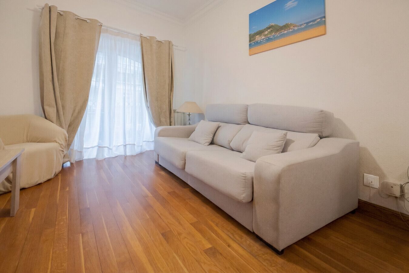 A living room with a sofa, a chair, wooden flooring, a lamp, and a painting on the wall.