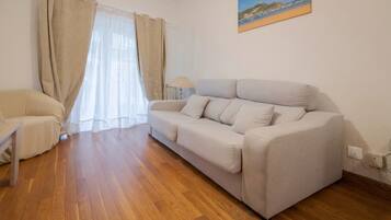 A living room with a sofa, a chair, wooden flooring, a lamp, and a painting on the wall.