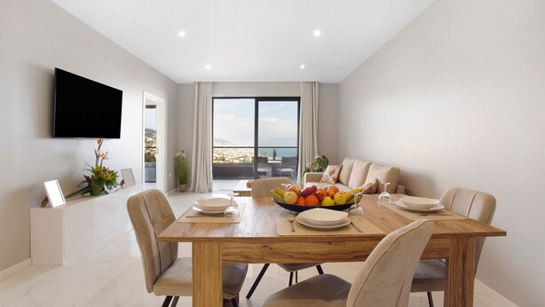 A modern dining area with a wooden table, beige chairs, a fruit bowl, and a flat-screen TV mounted on the wall. - Holiday Home 'Dream View' with Sea View, Private Terrace & Wi-Fi (Funchal)