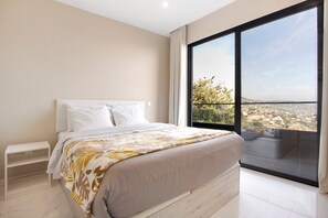 Een slaapkamer met een groot raam, een bed, een nachtkastje en uitzicht op een stadsgezicht.