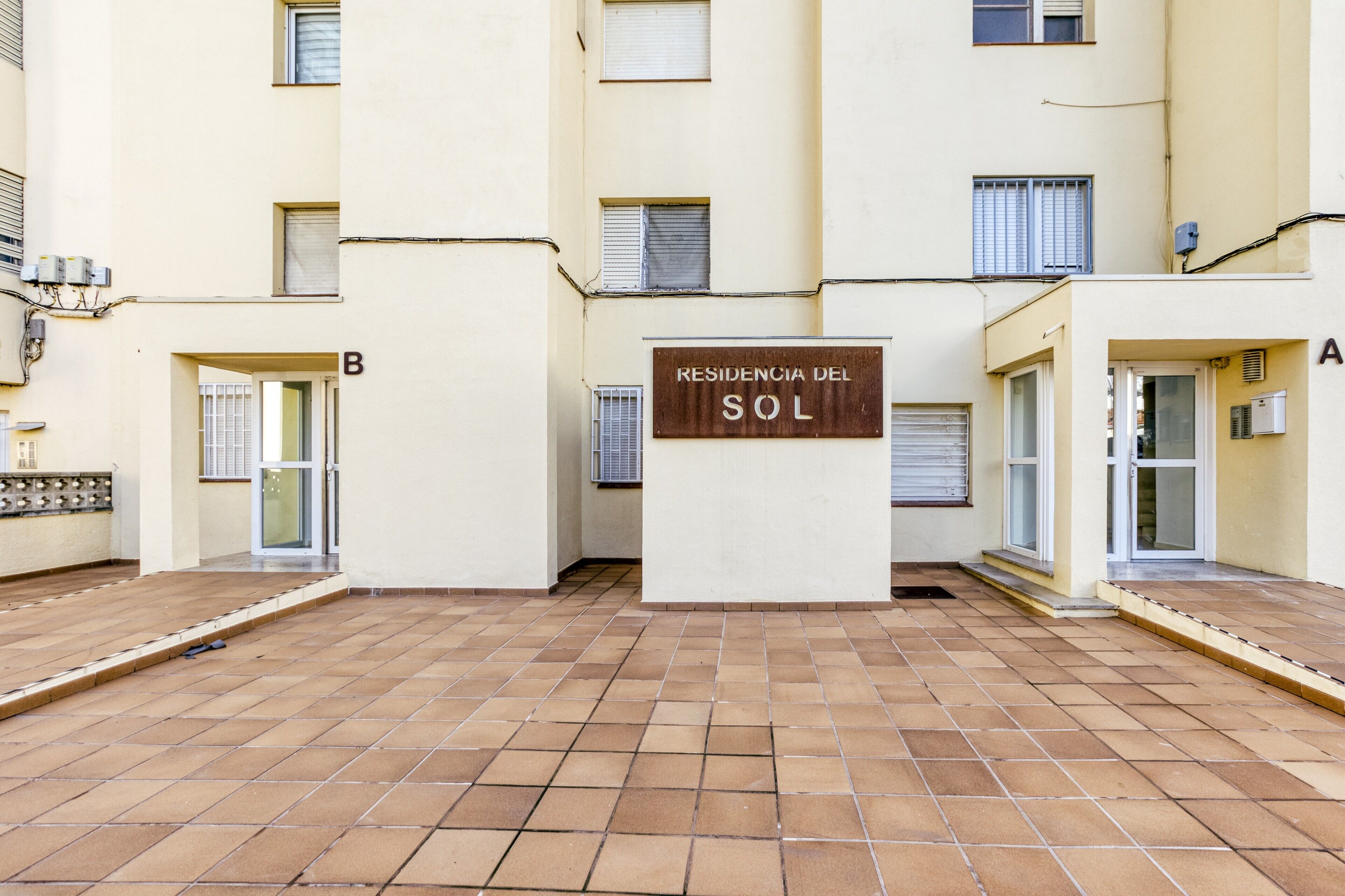Un immeuble résidentiel doté d’une cour dallée, d’une enseigne indiquant « Residencia del Sol » et d’une porte marquée de la lettre « B ».