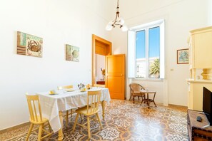 Comedor con una mesa lista para cuatro personas, una lámpara de araña, una ventana con vistas y una puerta que conduce a otra habitación.