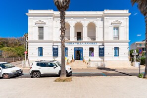 Un edificio blanco con entrada azul, identificado como 'Galleria Villa Ramirez', que presenta una fachada imponente con ventanas arqueadas y un balcón central.
