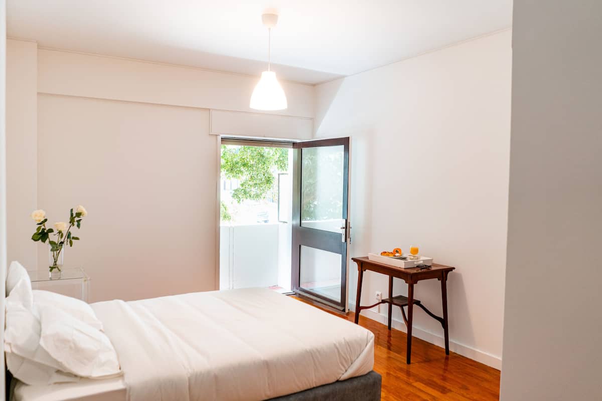 A bedroom with a bed, a nightstand with a vase of flowers, a table with a fruit bowl, and a sliding glass door leading to an outdoor area.