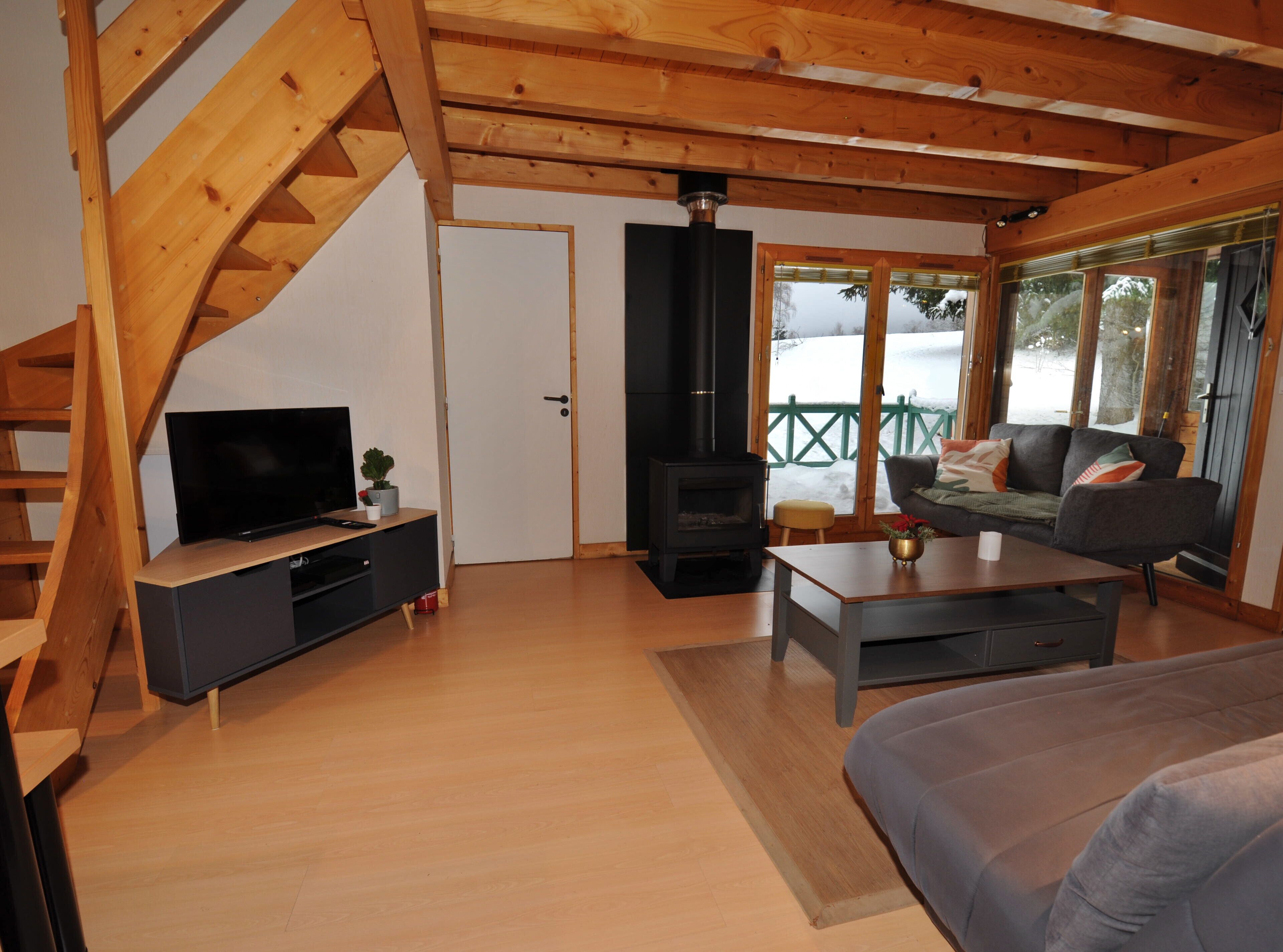 A cozy living room with a wooden staircase, a black wood-burning stove, a television on a wooden stand, and a sofa with cushions.