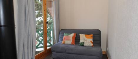 Une petite pièce avec un canapé gris, deux coussins, un réfrigérateur noir et une porte-fenêtre coulissante donnant sur un balcon.