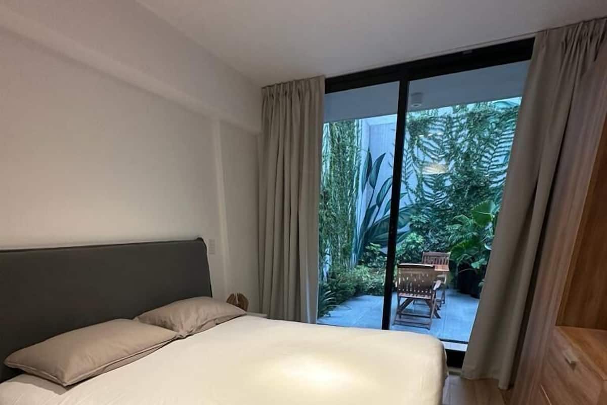 A modern bedroom with a large bed, wooden floor, and a view of a garden through a sliding glass door.