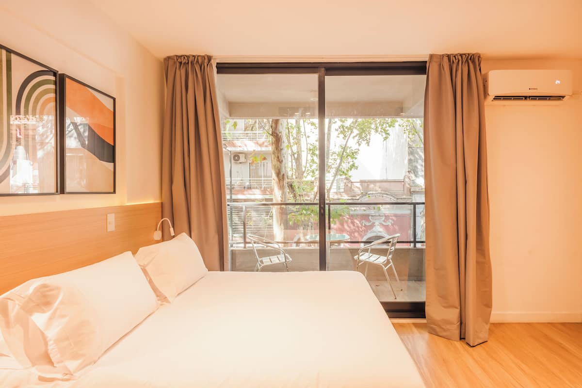 A hotel room with a bed, a large window with curtains, a balcony with chairs, and an air conditioning unit.