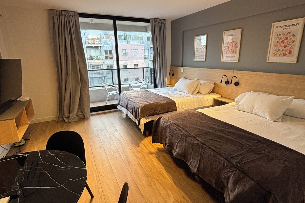 A hotel room with two beds, a desk, a chair, and a large window with a view of buildings.