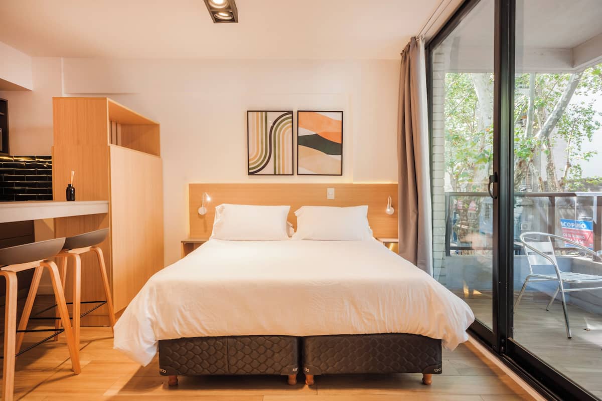 A modern hotel room with a large bed, a desk with two chairs, and a balcony with a view of trees and buildings.