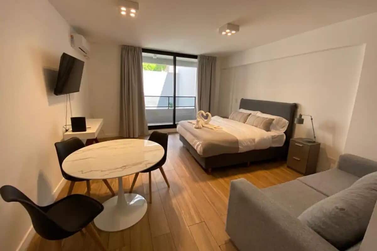 A modern hotel room with a bed, a sofa, a dining table with chairs, a TV, and a window with curtains.