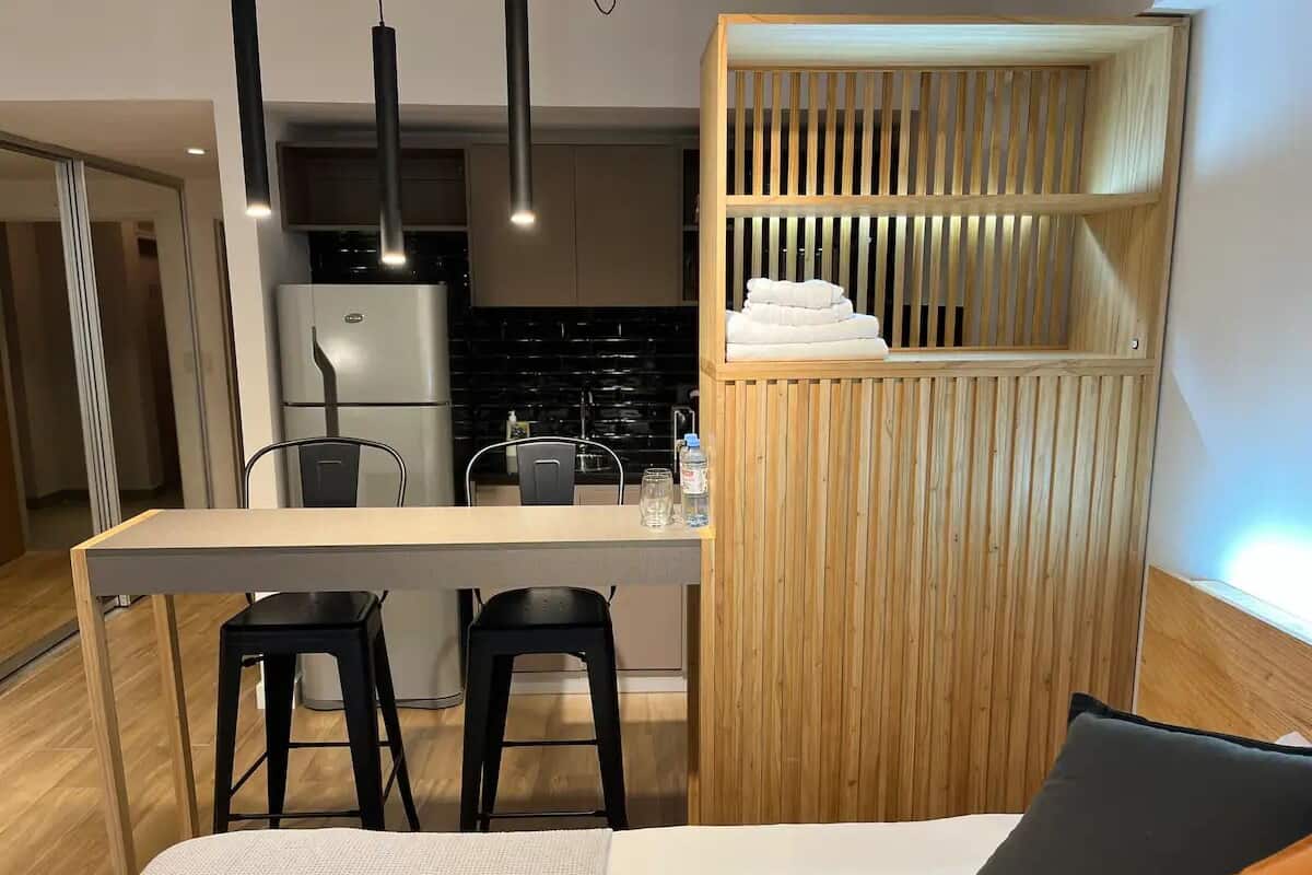 A compact hotel room with a bed, a small desk with two chairs, a kitchenette, and a wooden partition with towels.