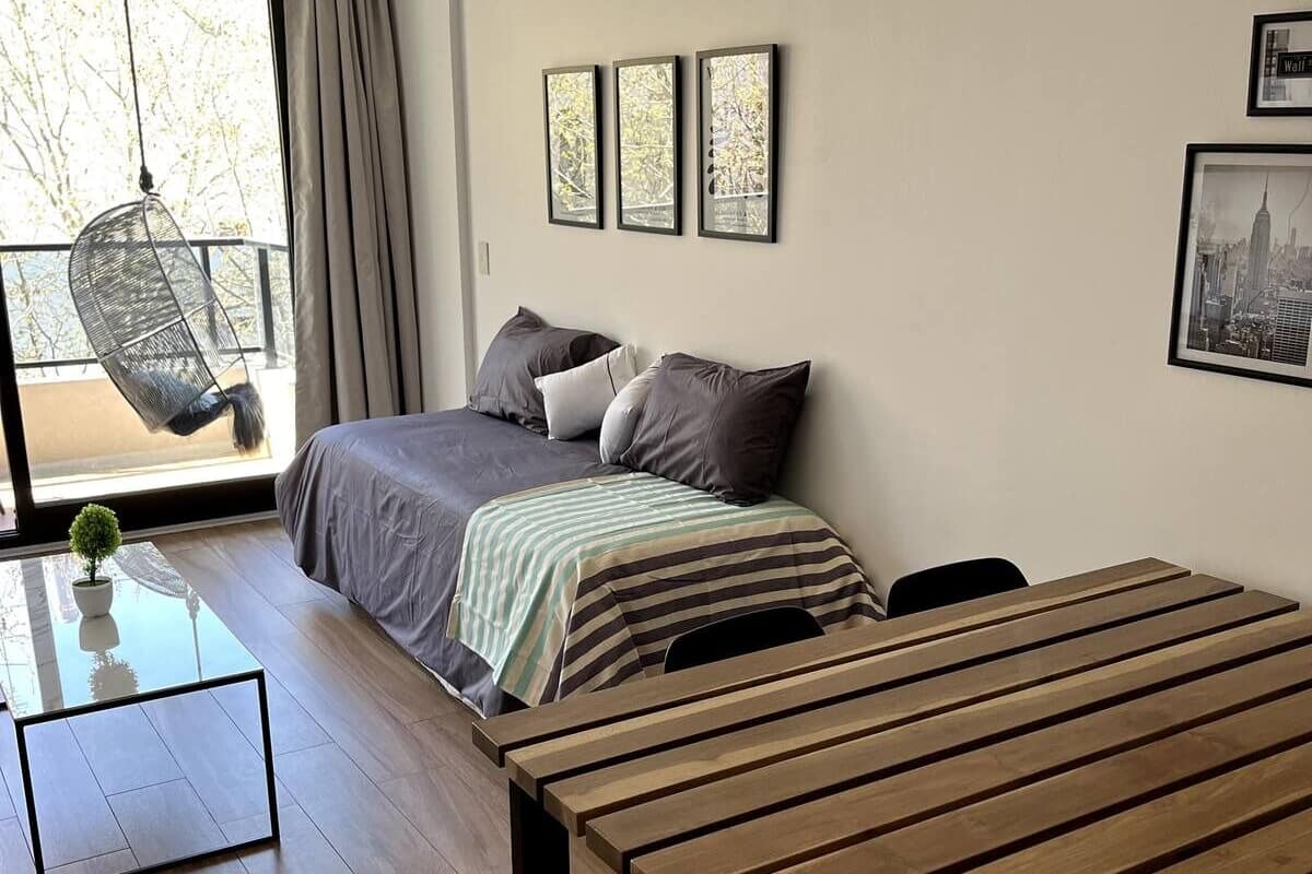 A modern bedroom with a large bed, a hanging chair by the window, and a wooden table.