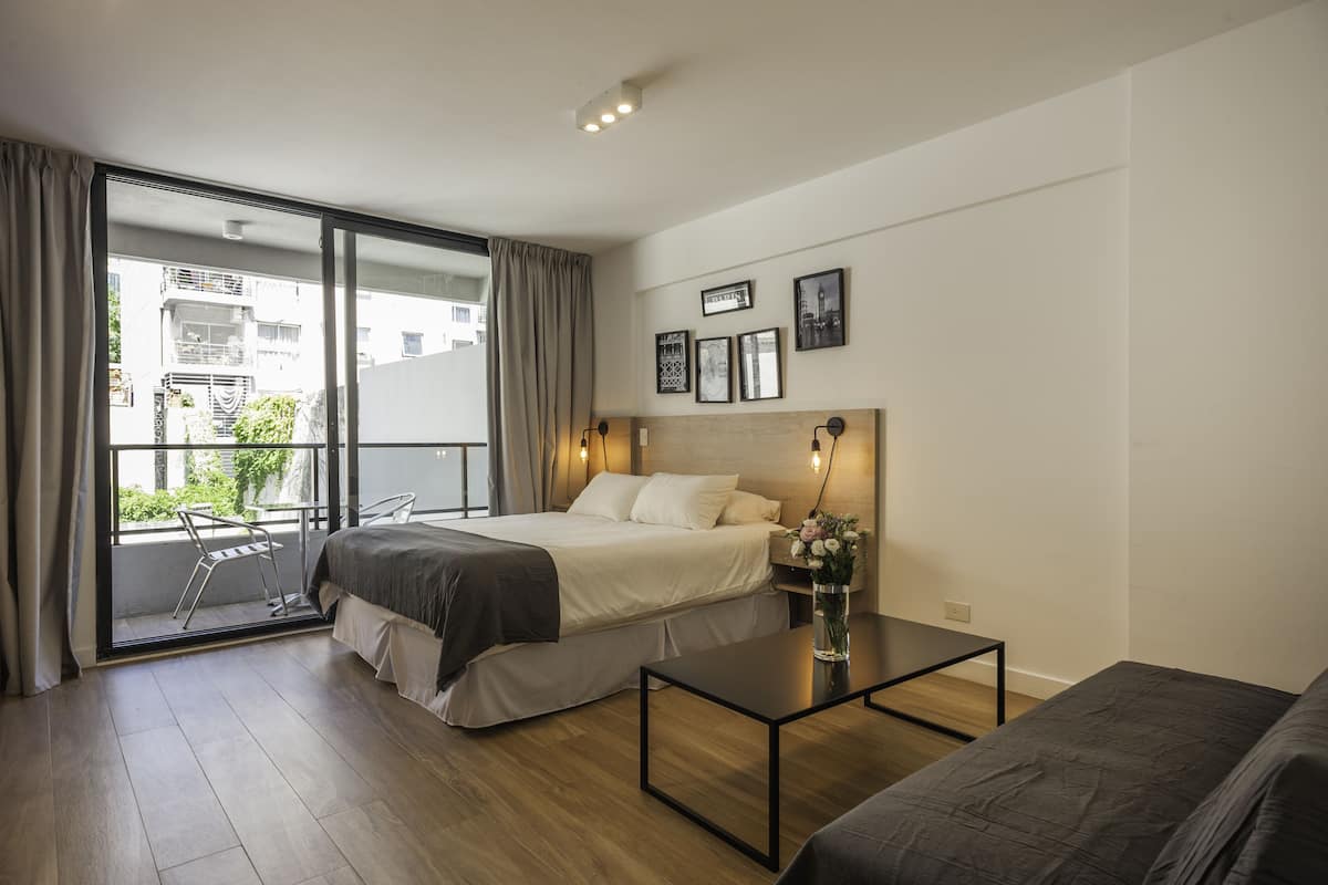 A modern hotel room with a bed, a small table, a chair, and a balcony with curtains.