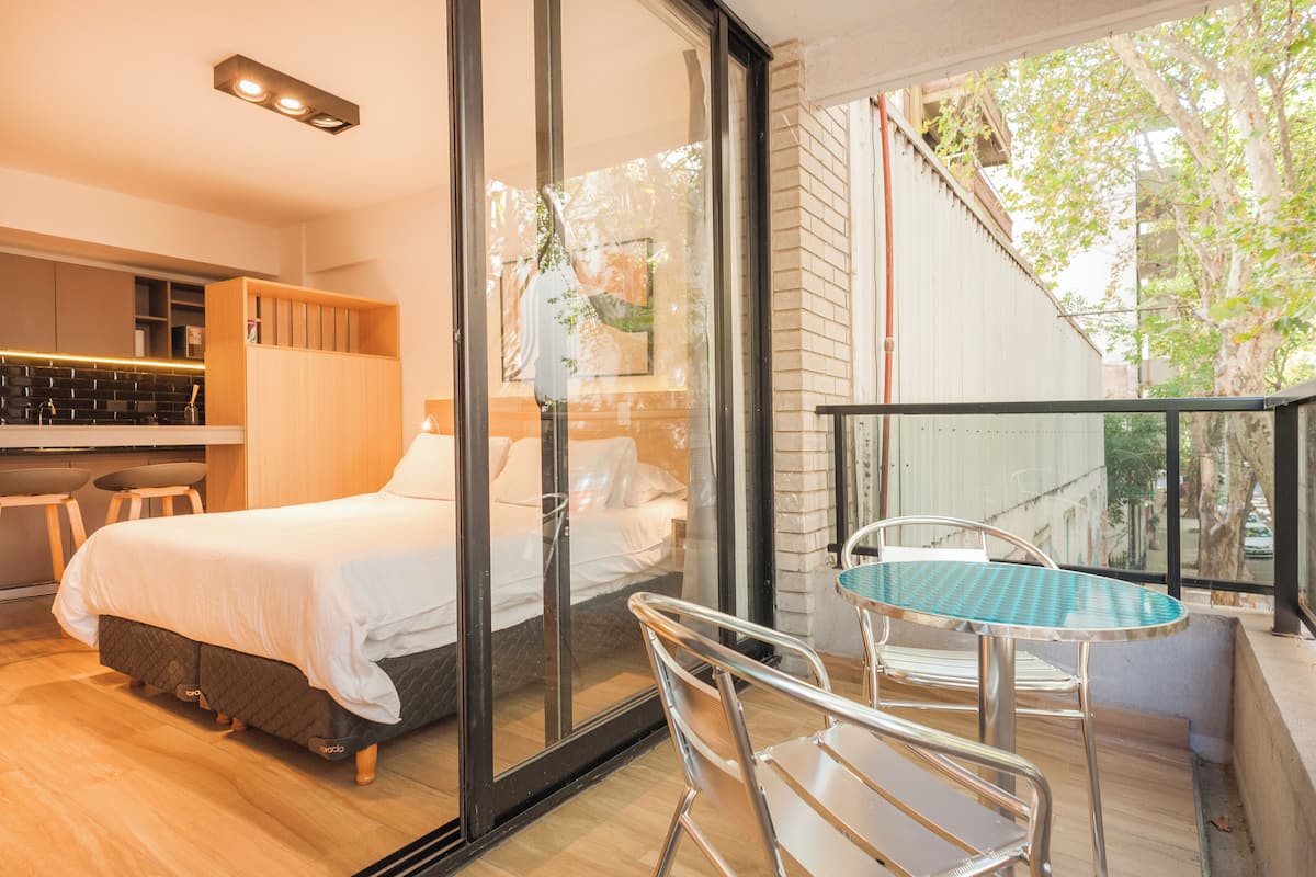 A modern bedroom with a glass door leading to a balcony, a small dining area, and a kitchen with a bar counter.