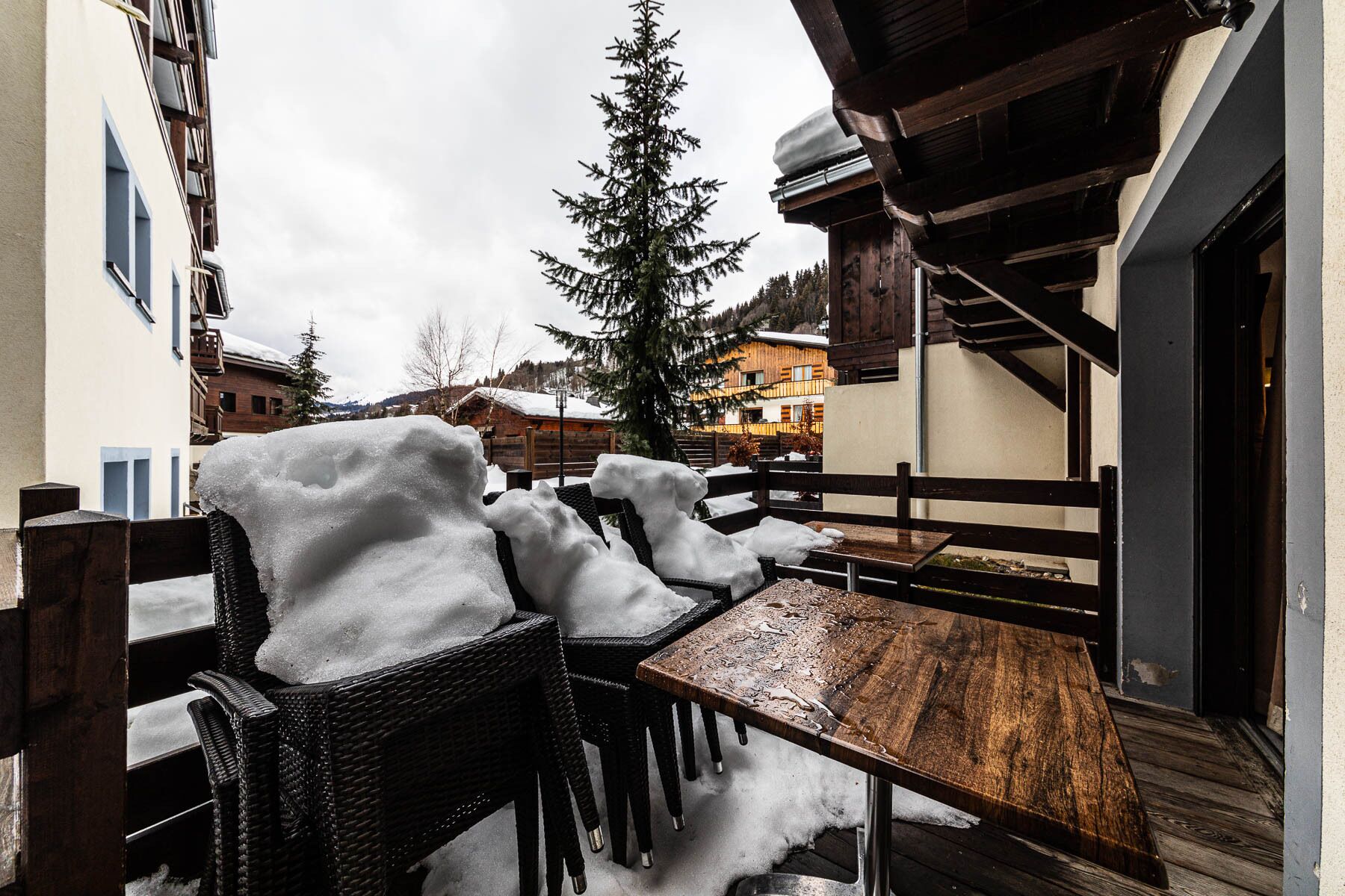 Uma área externa coberta de neve com cadeiras de vime e uma mesa de madeira, cercada por edifícios e uma árvore com neve.