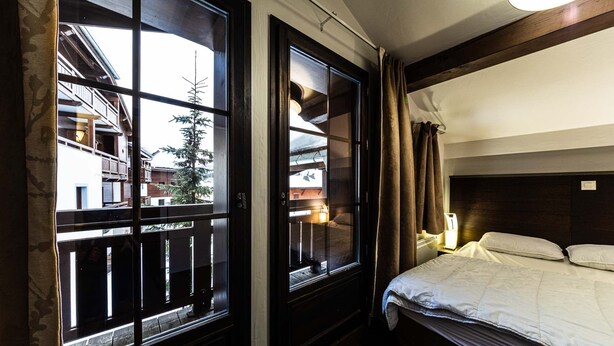 Habitación de hotel con cama, mesita de noche, lámpara y dos ventanas con cortinas.