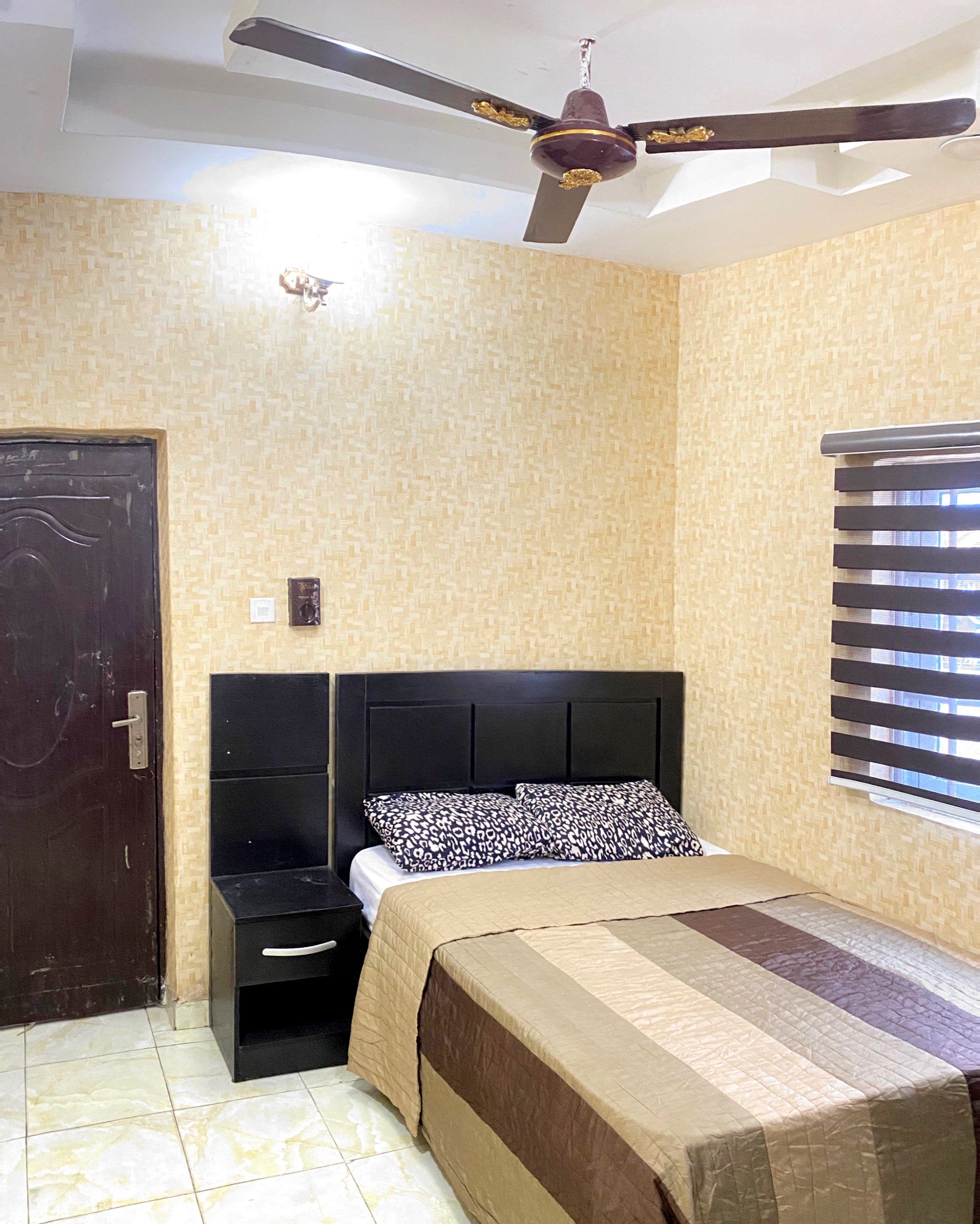 A bedroom with a bed, a nightstand, a ceiling fan, a window with blinds, and a door.