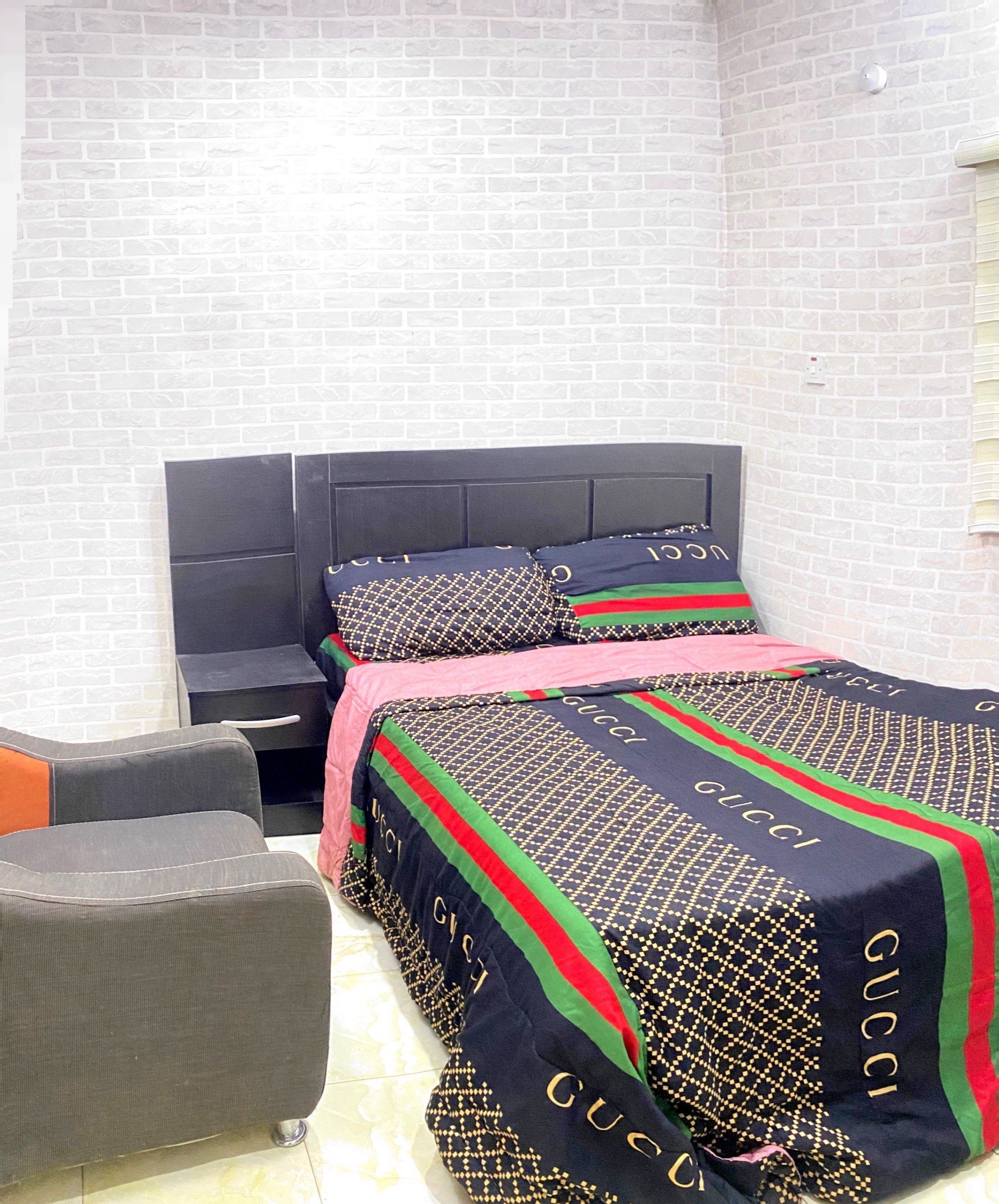 Habitación de hotel con pared de ladrillo, cama con cubrecama de Gucci, sillón gris y mesita de noche.