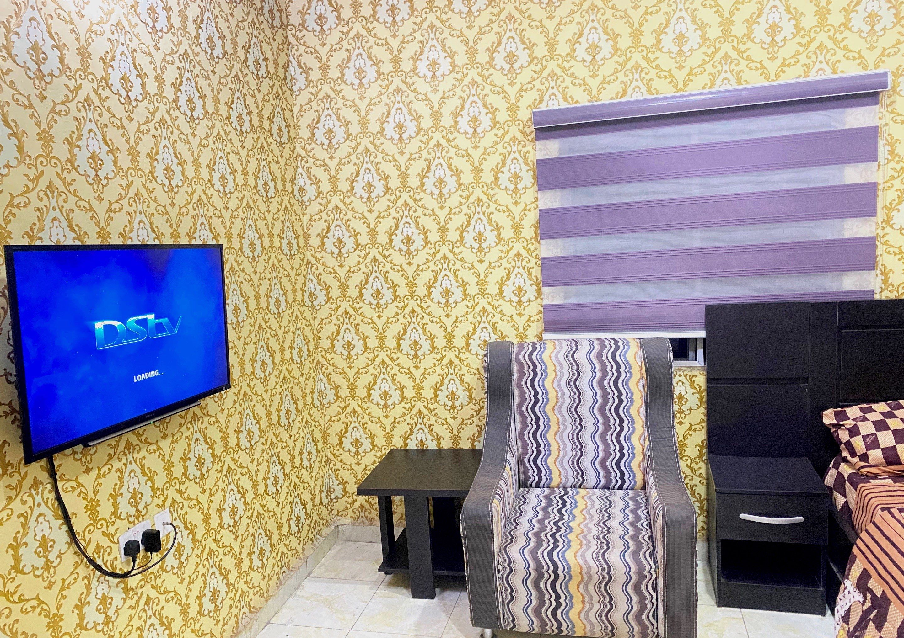 Ein Zimmer mit gemustertem Wandpapier, einem Fernseher, der das 'DSN'-Logo zeigt, einem gemusterten Sessel, einem kleinen Tisch und einem Bett mit karierter Decke.