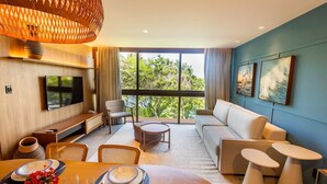Un salon moderne doté d’un grand téléviseur, de sièges confortables, d’un coin repas et de grandes fenêtres offrant une vue sur la verdure.