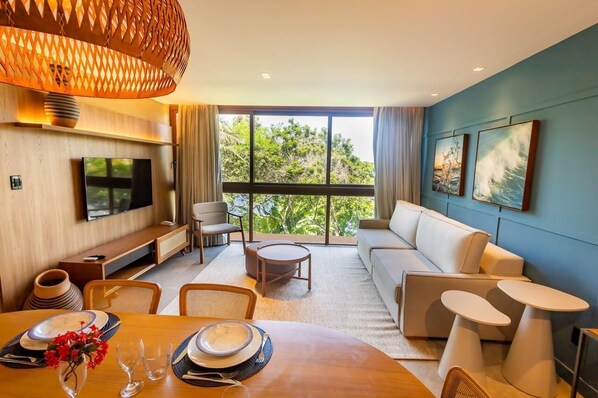 A modern living room with a large TV, comfortable seating, a dining area, and large windows offering a view of greenery. - Guarajuba Suites Premium Boutique Hotel (Camaçari)