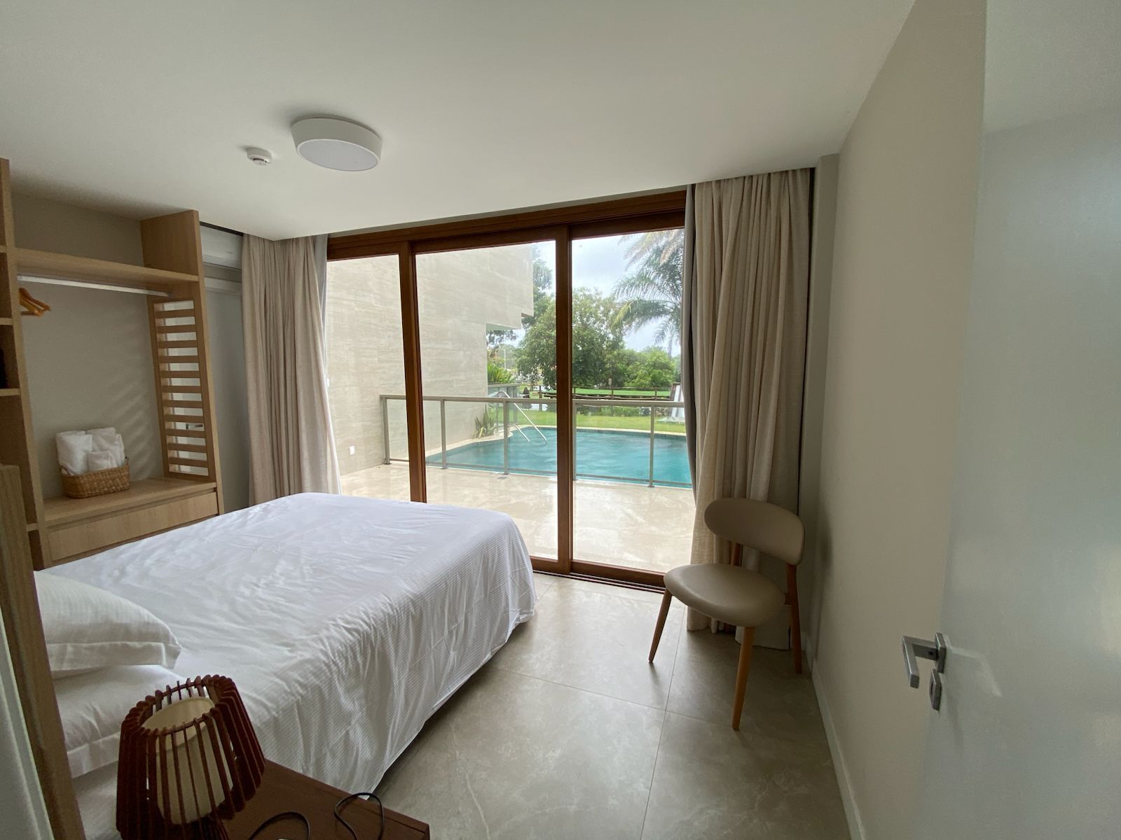 A hotel room with a bed, a chair, a small table, a window with a view of a swimming pool, and a balcony.