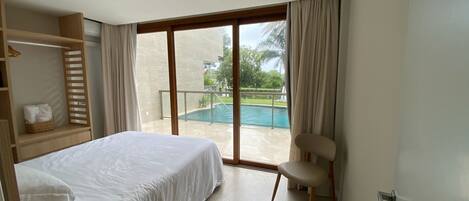 A hotel room with a bed, a chair, a small table, a window with a view of a swimming pool, and a balcony.