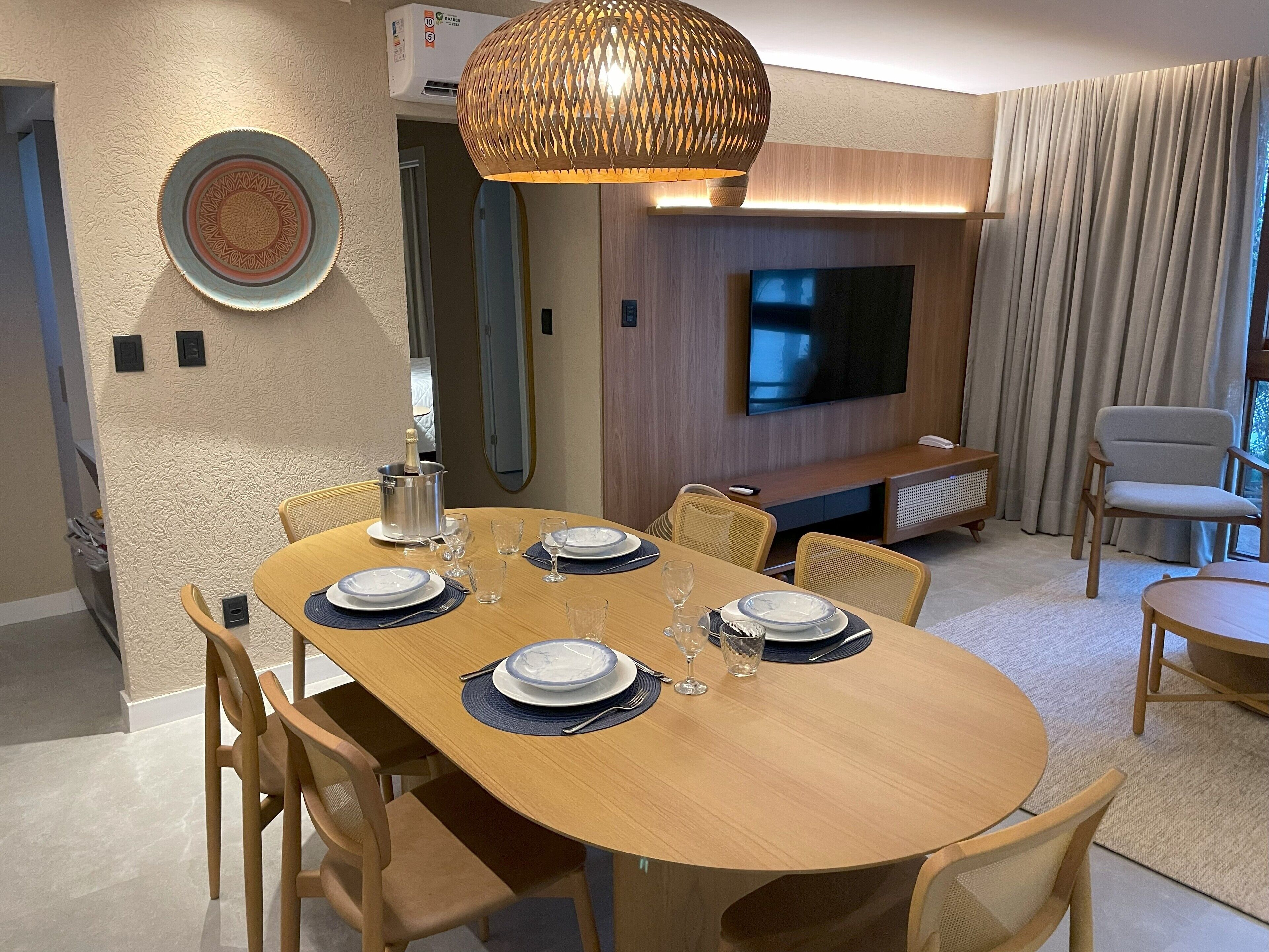 Un comedor moderno con una mesa de madera dispuesta para cuatro comensales, una lámpara colgante de fibras naturales, un televisor de pantalla plana y un conjunto de asientos cómodos.