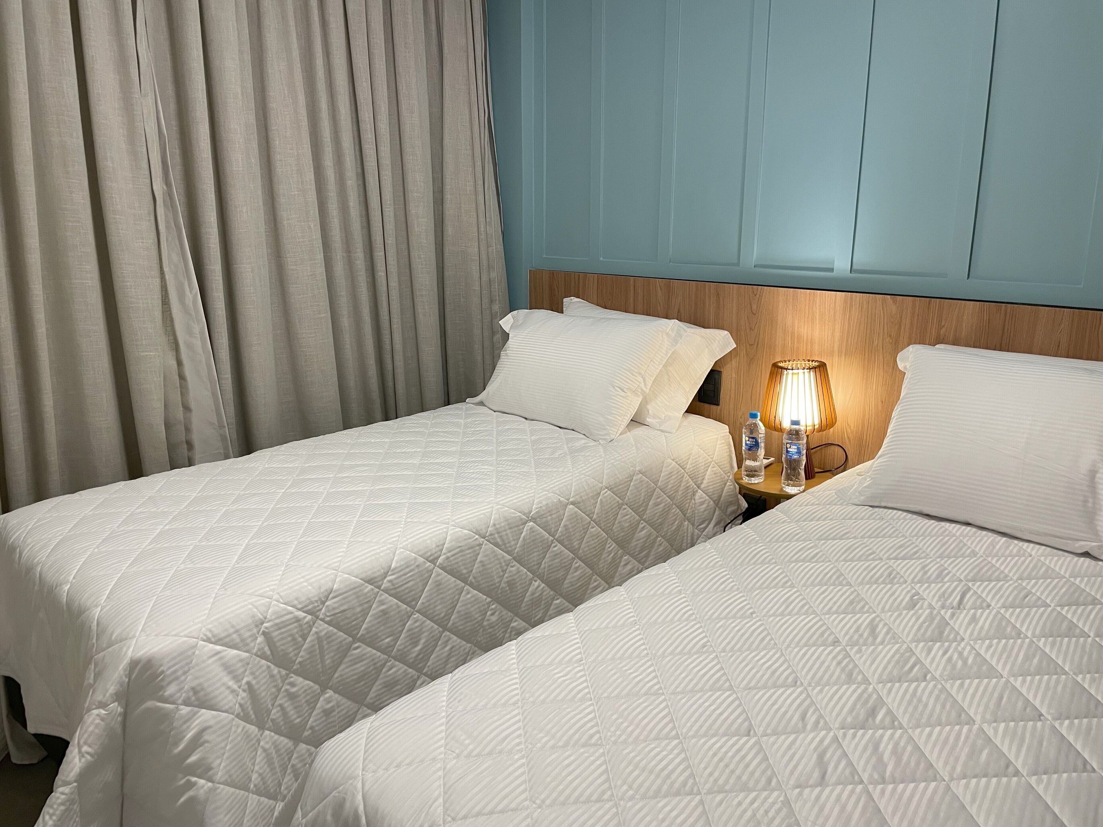 Une chambre d’hôtel avec deux lits, une tête de lit en bois, une table de chevet avec une lampe et une fenêtre avec des rideaux.