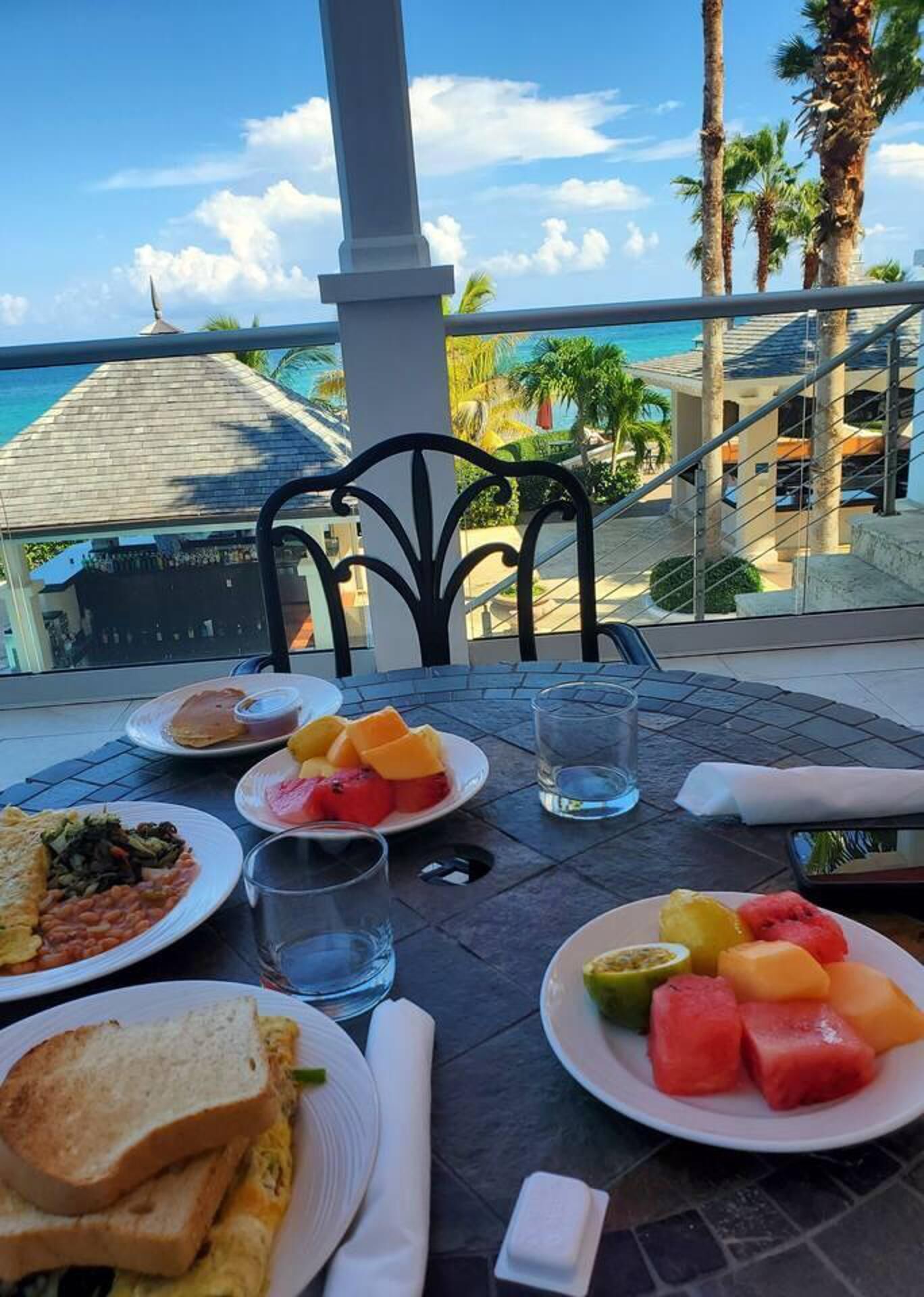 Una mesa servida con alimentos para el desayuno, como tostadas, huevos, frijoles y fruta, con vistas a un paisaje pintoresco con palmeras y un edificio.