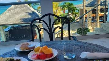 Una mesa servida con alimentos para el desayuno, como tostadas, huevos, frijoles y fruta, con vistas a un paisaje pintoresco con palmeras y un edificio.