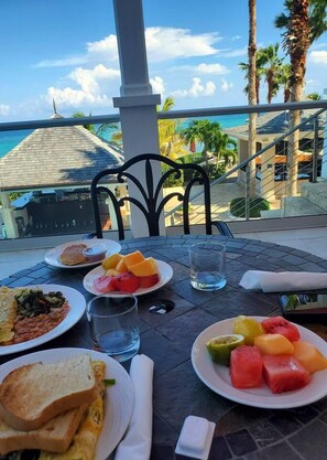 Une table dressée pour le petit-déjeuner, avec des toasts, des œufs, des haricots et des fruits, donnant sur un paysage pittoresque avec des palmiers et un bâtiment.