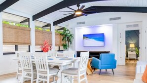 Un espace repas moderne avec une table et des chaises blanches, un fauteuil bleu et un téléviseur fixé au mur.