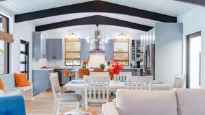 Une cuisine moderne avec des meubles blancs, un coin repas avec des chaises blanches et une grande fenêtre équipée de stores.