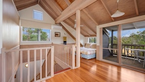A spacious room with a wooden floor, a bed, a desk, and a large window offering a view of the outdoors. - Wollondilly - Mansfield - Sleeps 10 (Goughs Bay)