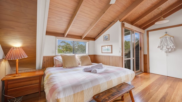 A bedroom with a wooden ceiling, a bed with a checkered bedspread, a bedside lamp, a wooden bench, and a window with a view of trees. - Wollondilly - Mansfield - Sleeps 10 (Goughs Bay)