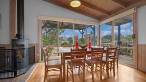 A dining area with a large table and chairs, a fireplace, and large windows offering a view of the outdoors. - Wollondilly - Mansfield - Sleeps 10 (Goughs Bay)