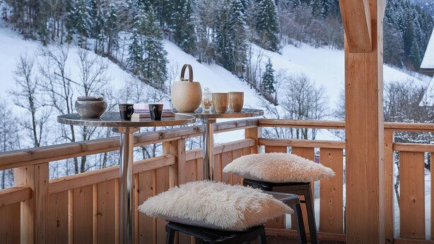 Um deck de madeira com uma mesa posta para o chá, rodeado por árvores cobertas de neve e uma montanha ao fundo.