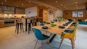 Uma área de refeições moderna com uma mesa de madeira comprida, cadeiras azuis e amarelas, e uma cozinha ao fundo.