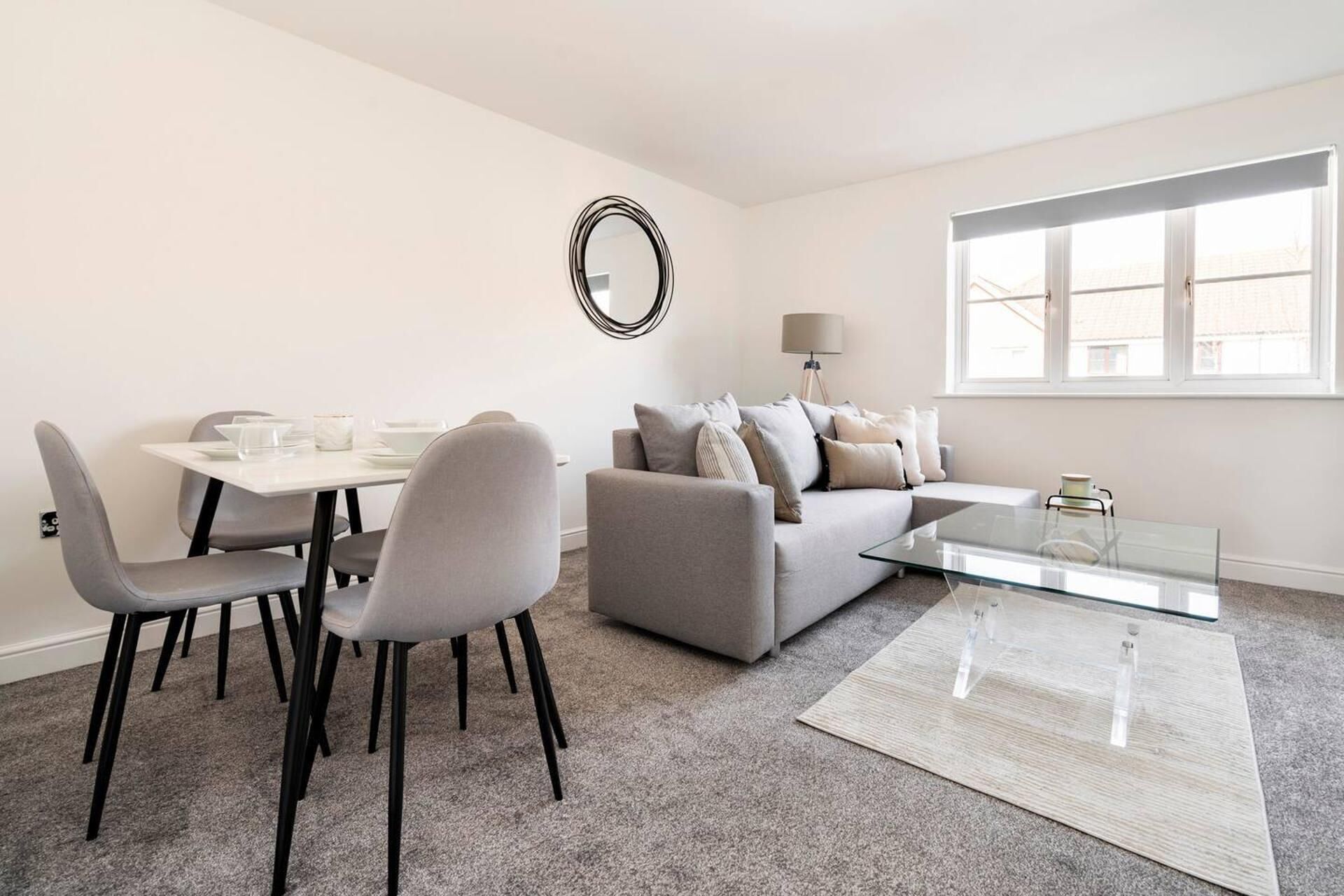 Ein modernes Wohnzimmer mit einem grauen Sofa, einem Glastisch und einem Essbereich mit weißem Tisch und Stühlen.