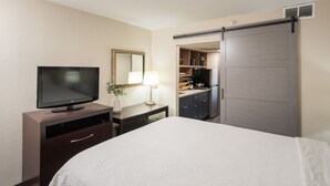 A hotel room with a bed, a TV, a desk with a lamp, a wardrobe, and a kitchenette. - South Country Suites (Mesa)