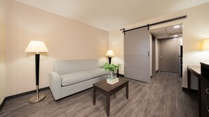 A modern hotel room with a grey sofa, two floor lamps, a wooden coffee table with a book and a vase, and a sliding door leading to a bathroom. - South Country Suites (Mesa)