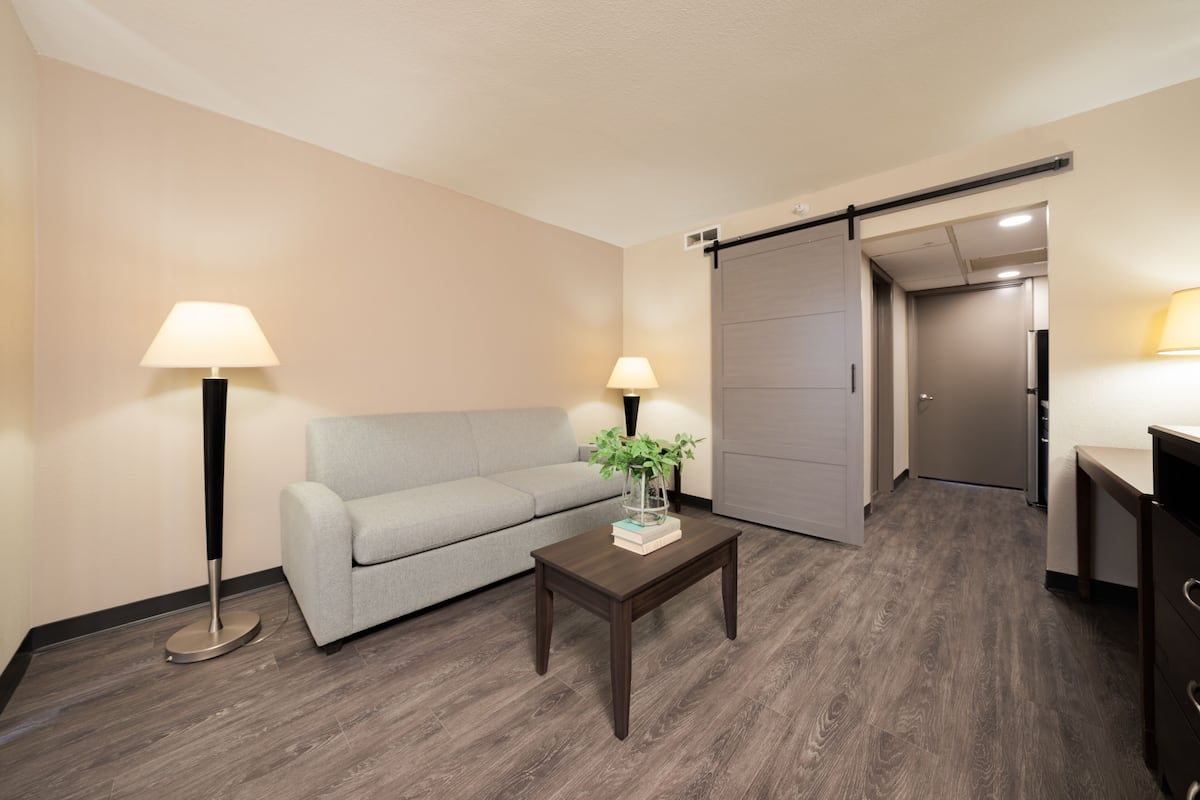A modern hotel room with a grey sofa, two floor lamps, a wooden coffee table with a book and a vase, and a sliding door leading to a bathroom.